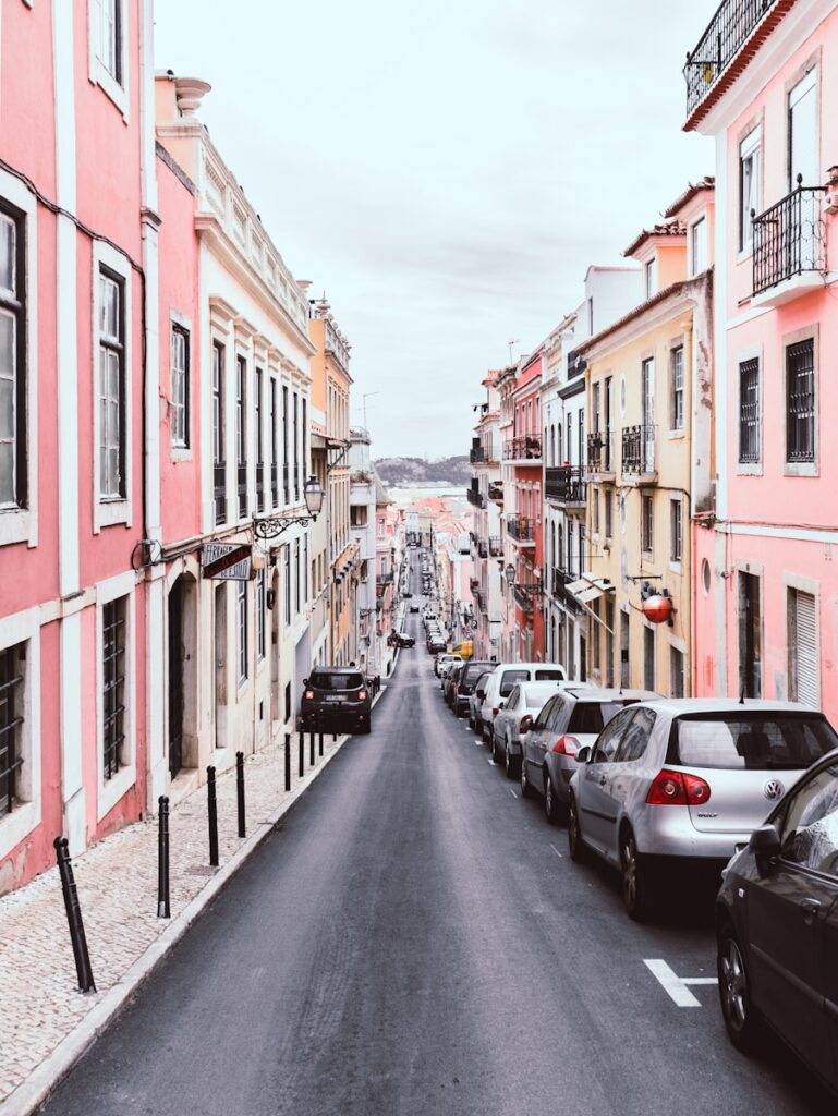 Lisbon
line of vehicles on the right side of a 2-way road in the middle of pink and yellow buildings during day