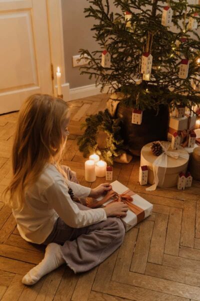 Young girl opening a gift under a decorated Christmas tree with candles and ornaments nearby.