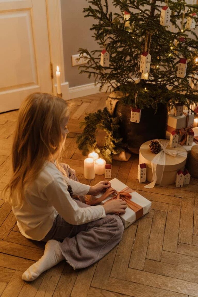 Young girl opening a gift under a decorated Christmas tree with candles and ornaments nearby.