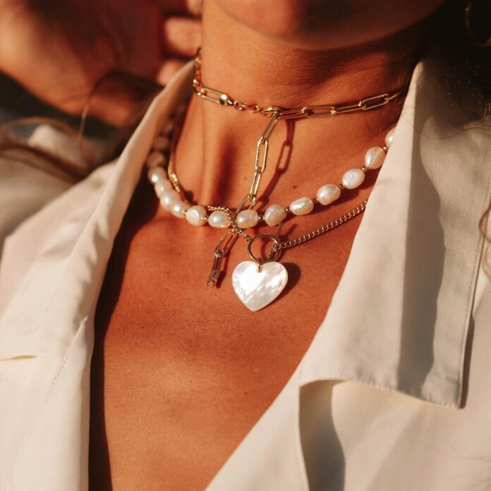 layer necklace - Close-up of elegant layered necklaces featuring pearls and gold chains on sunlit skin.