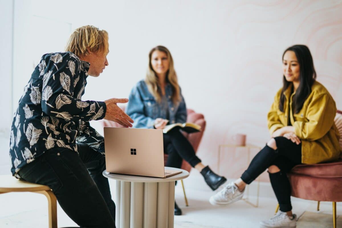 3 Proven Principles to Boost Team Creativity - man in black and white floral long sleeve shirt sitting and showing something on a microsoft laptop to 2 women