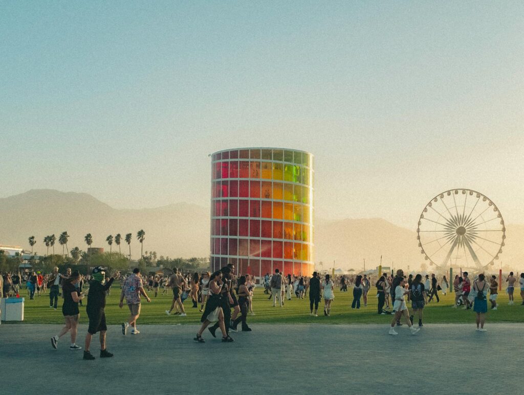 Coachella, People enjoying a lively music festival with a colorful tower and Ferris wheel backdrop.