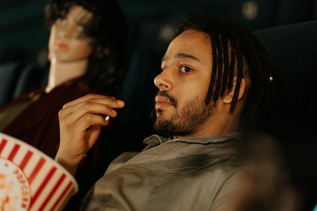 A man with dreadlocks enjoys popcorn in a cinema, sitting next to a mannequin.