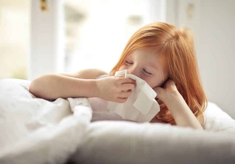 flu - Redheaded child resting in bed, wiping nose with tissue, unwell with flu-like symptoms.